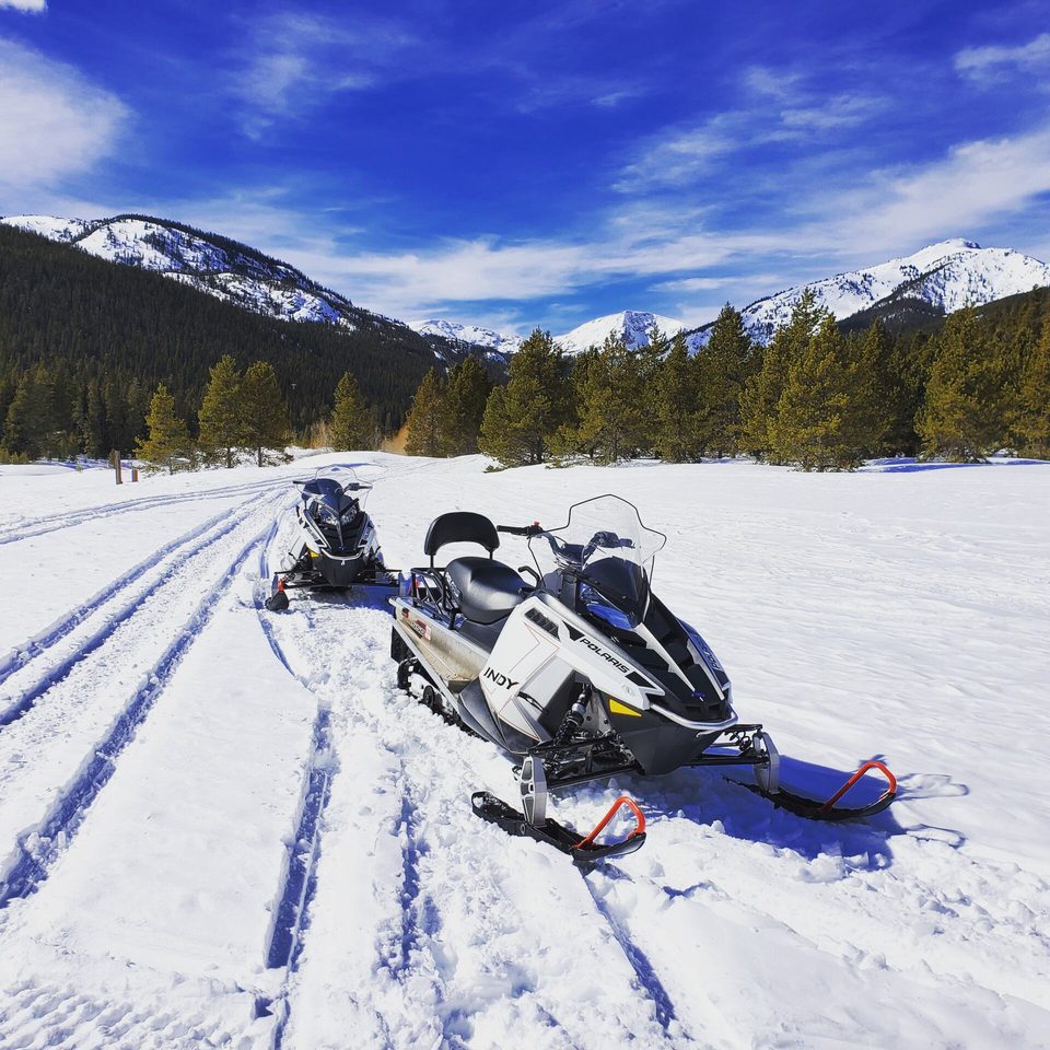 Two snowmobiles parked on a groomed trail under a deep blue sky, snowy peaks behind the trees