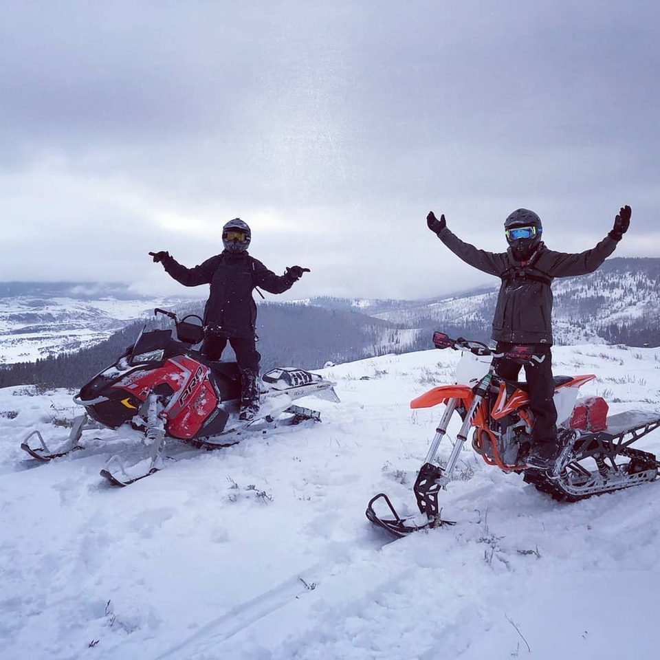 Two riders standing arms-out on a snowy ridge with valleys and clouds behind